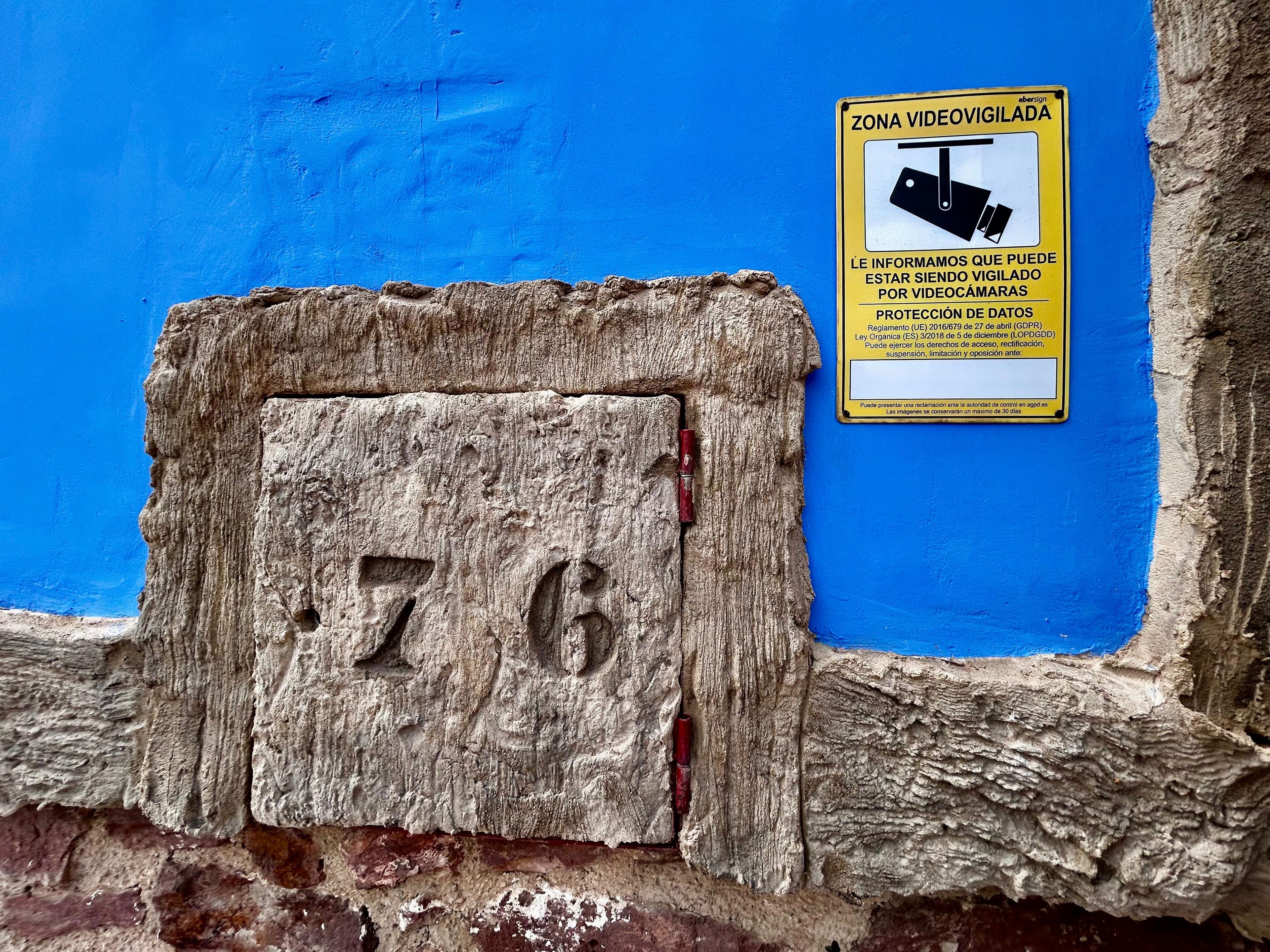 Un mur bleu sur lequel est apposée une pancarte jaune indiquant une zone sous vidéosurveillance, ainsi qu’une plaque en bois portant le numéro 76.
