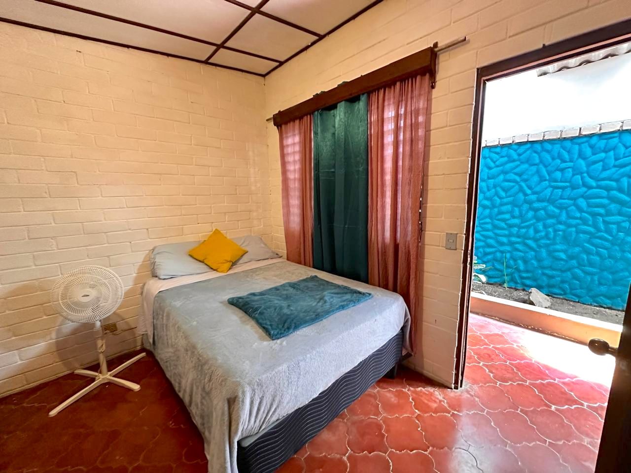 Une pièce avec un lit, un ventilateur sur pied et une vue sur un mur bleu à l’extérieur.