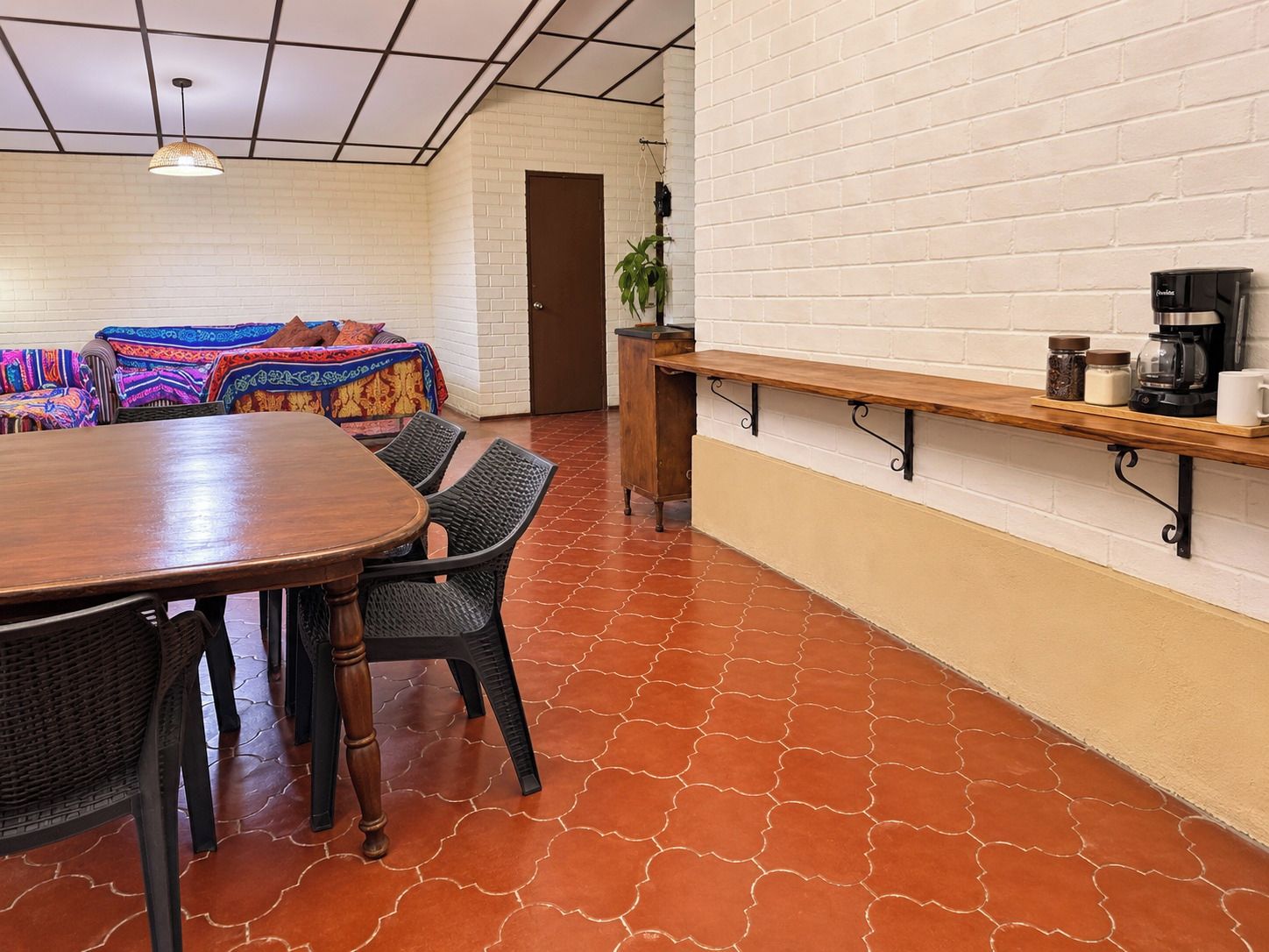 Un coin repas avec une table et des chaises en bois, un banc avec une machine à café, et un mur en briques blanches.