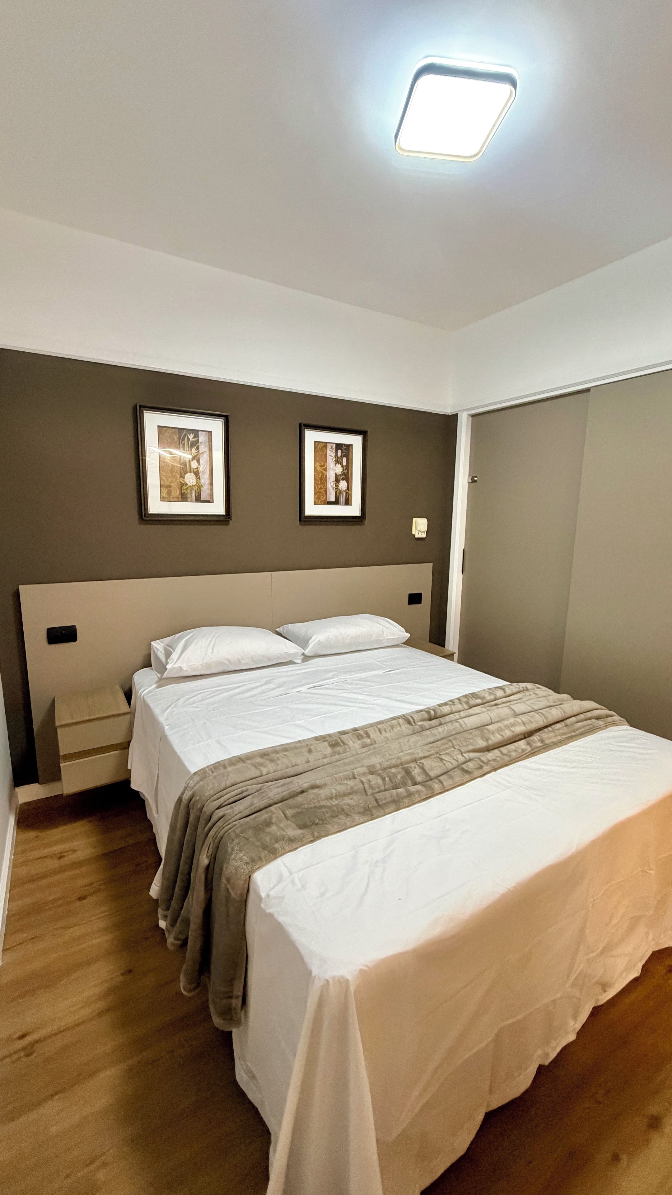 Een hotelkamer met een bed, twee foto’s in lijsten aan de muur, een nachtkastje en een lamp boven het bed.