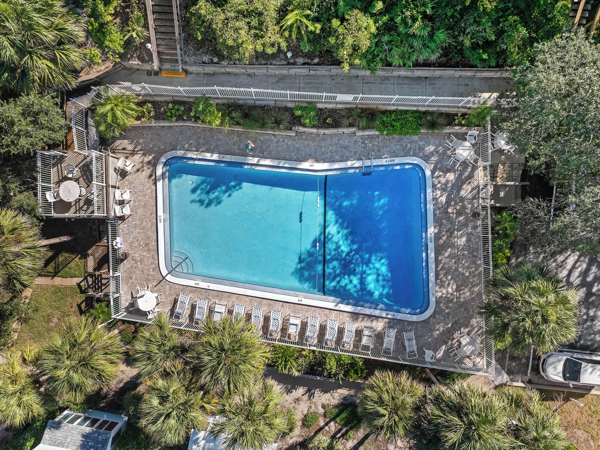 An aerial view of a rectangular swimming pool with a surrounding deck, lounge chairs, and a small bridge.