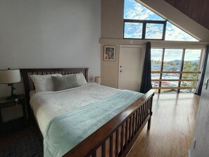 Ein Schlafzimmer mit einem großen Bett, einem Nachttisch, einer Lampe, einer Tür und Blick auf einen Yachthafen durch ein großes Fenster.