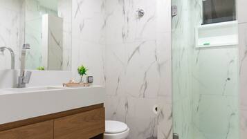 A modern bathroom with a marble backsplash, a wooden vanity, a toilet, a shower with a glass enclosure, and a blue trash bin.