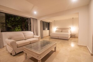 Una sala de estar moderna con un sofá color beige, una mesa de centro de vidrio y una cama con cabecera.