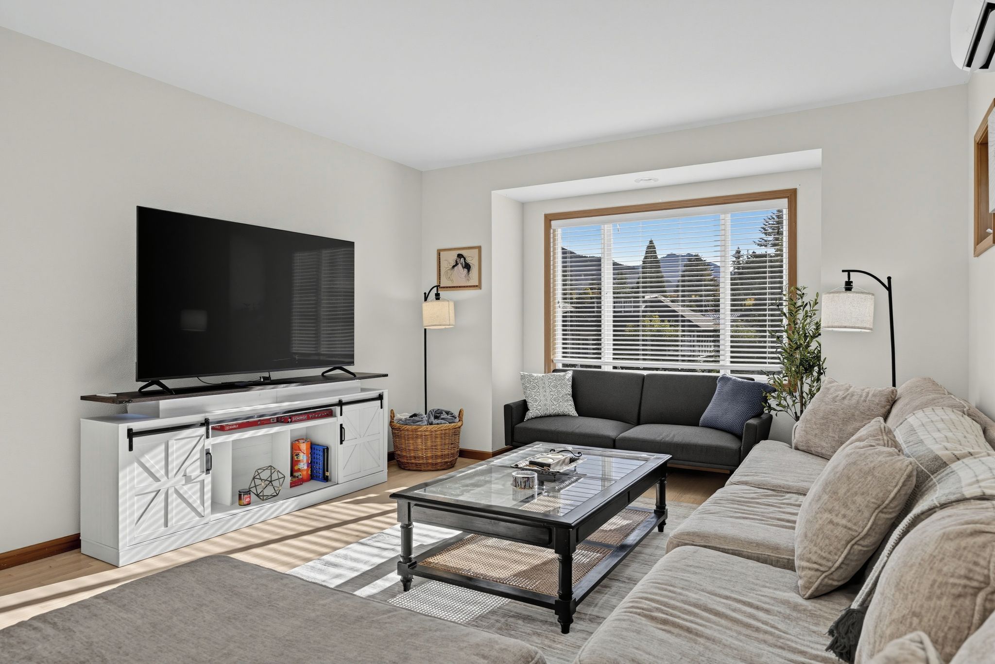 A modern living room with a flat-screen TV, a sofa, a coffee table, and a large window with blinds.