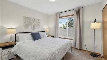 A bedroom with a bed, bedside tables, a lamp, a macrame wall hanging, and a window with curtains.