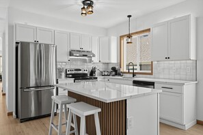 A modern kitchen with white cabinetry, stainless steel appliances, a central island with stools, and a window with a brick pattern view. - Spacious 3-bedroom North Bend with AC (North Bend)
