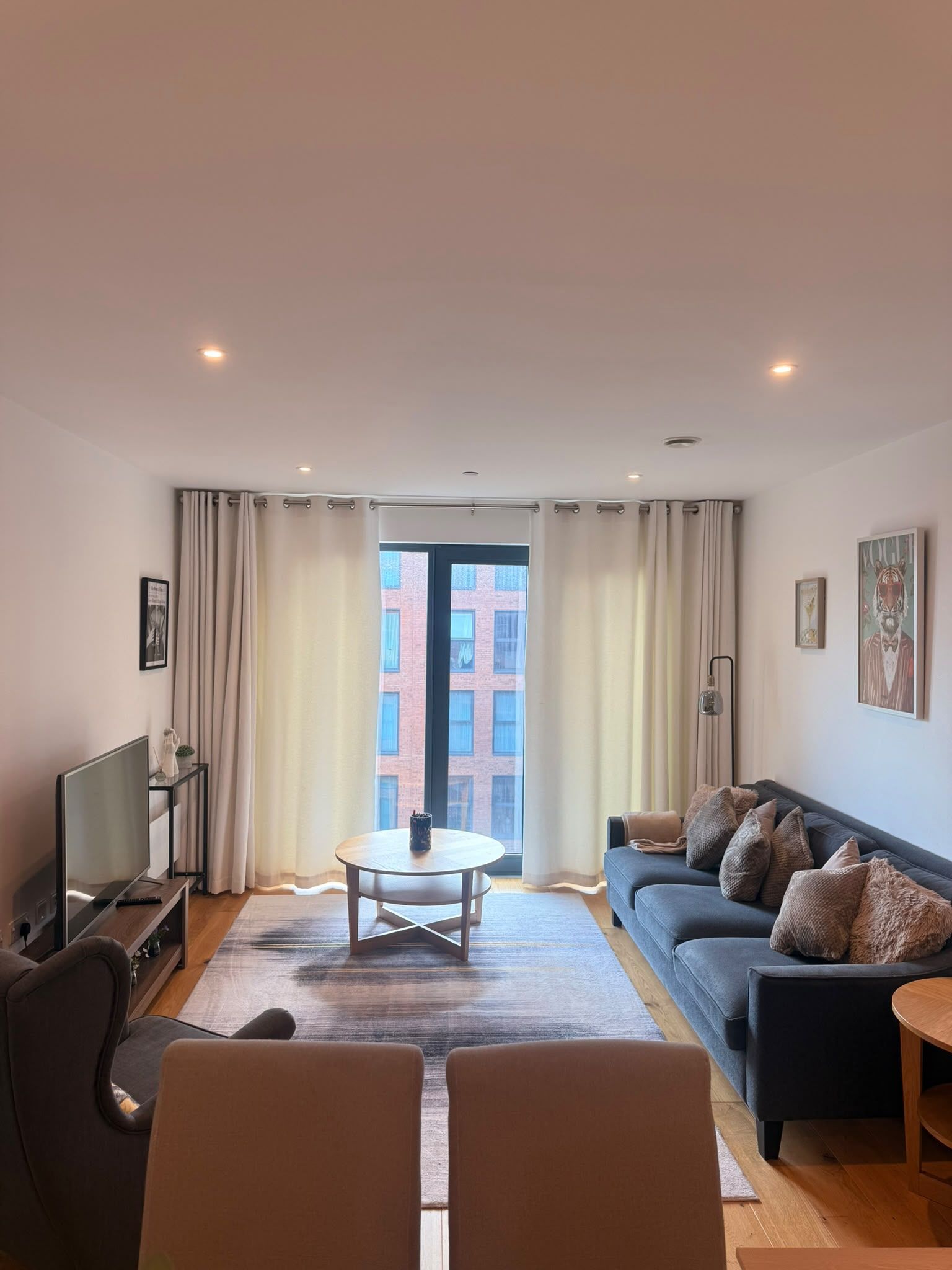A modern living room with a sofa, a TV, a coffee table, and a view of a building through a large window.