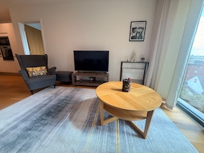 Un salon moderne avec un canapé, un téléviseur, une table basse et une vue sur la ville grâce à une grande fenêtre.