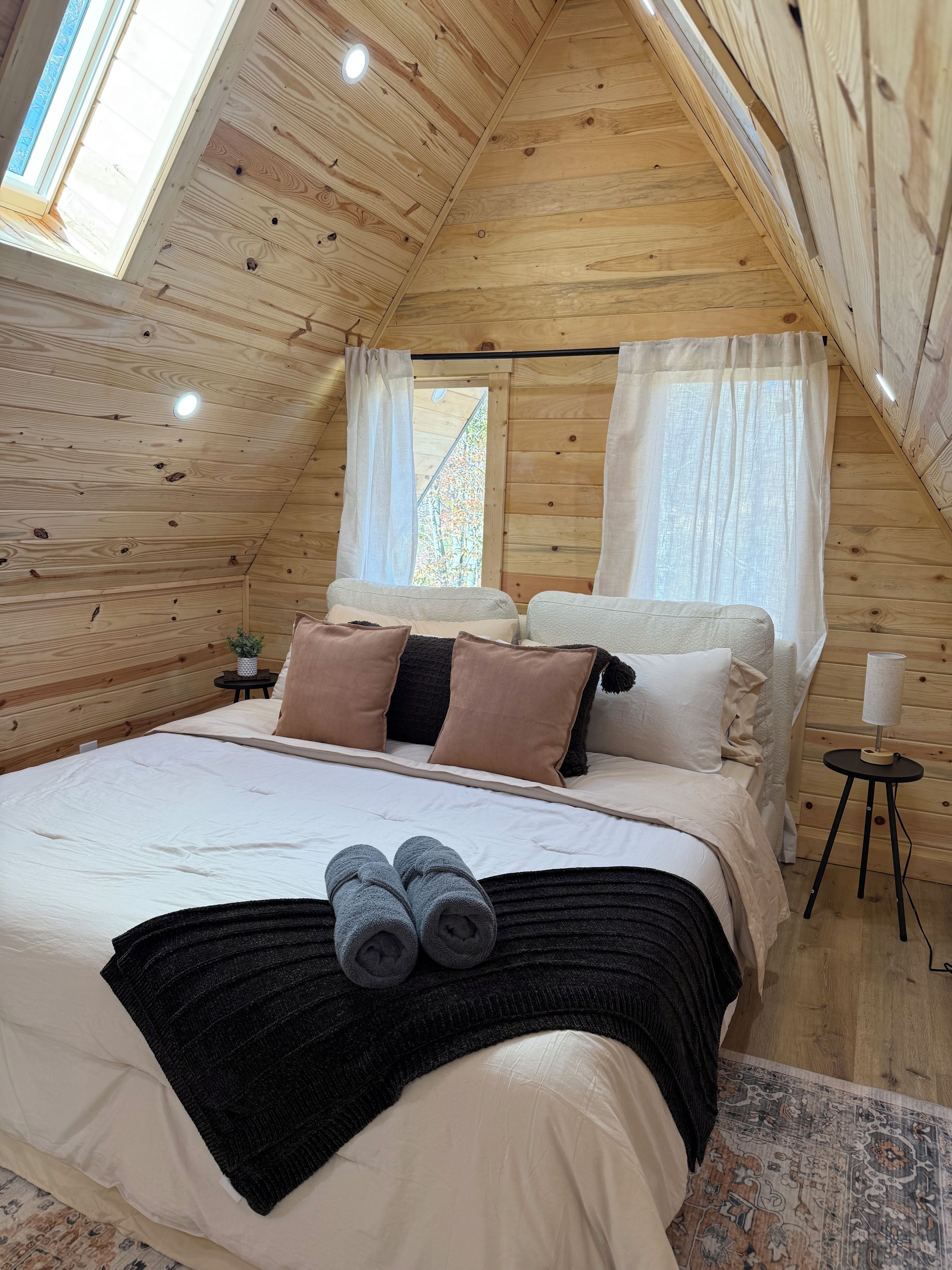 A cozy bedroom with a sloped ceiling, a bed with multiple pillows, a bedside table with a lamp, and a window with curtains.