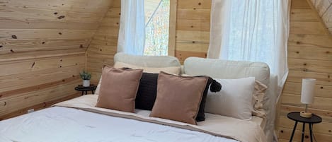 A cozy bedroom with a sloped ceiling, a bed with multiple pillows, a bedside table with a lamp, and a window with curtains.
