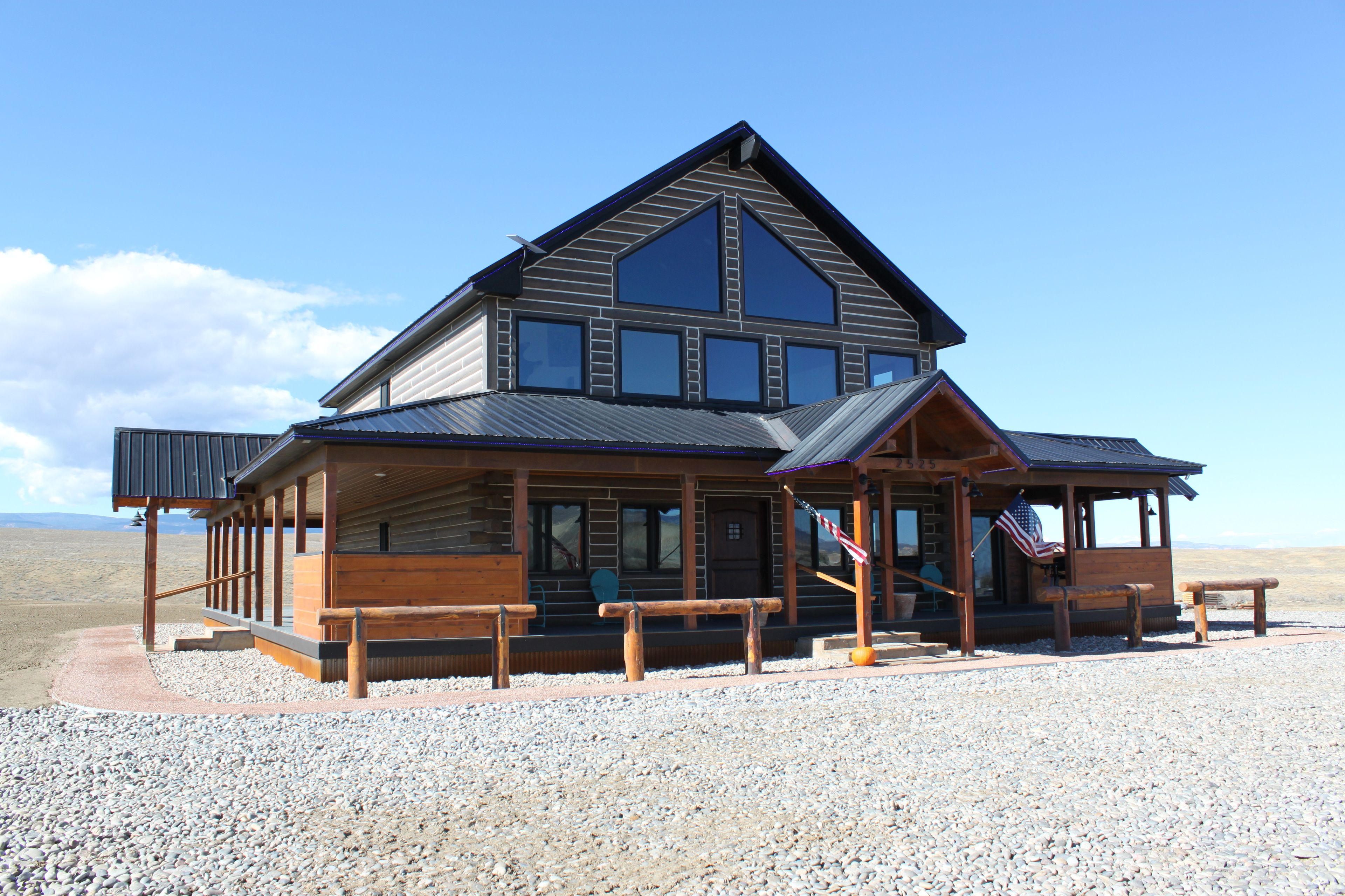 A two-story log cabin with a prominent gable roof, a porch with wooden railings, and a flagpole with an American flag.