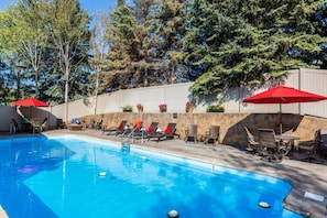 Piscina com cadeiras de descanso e guarda-sóis, cercada por árvores e um muro de pedra.