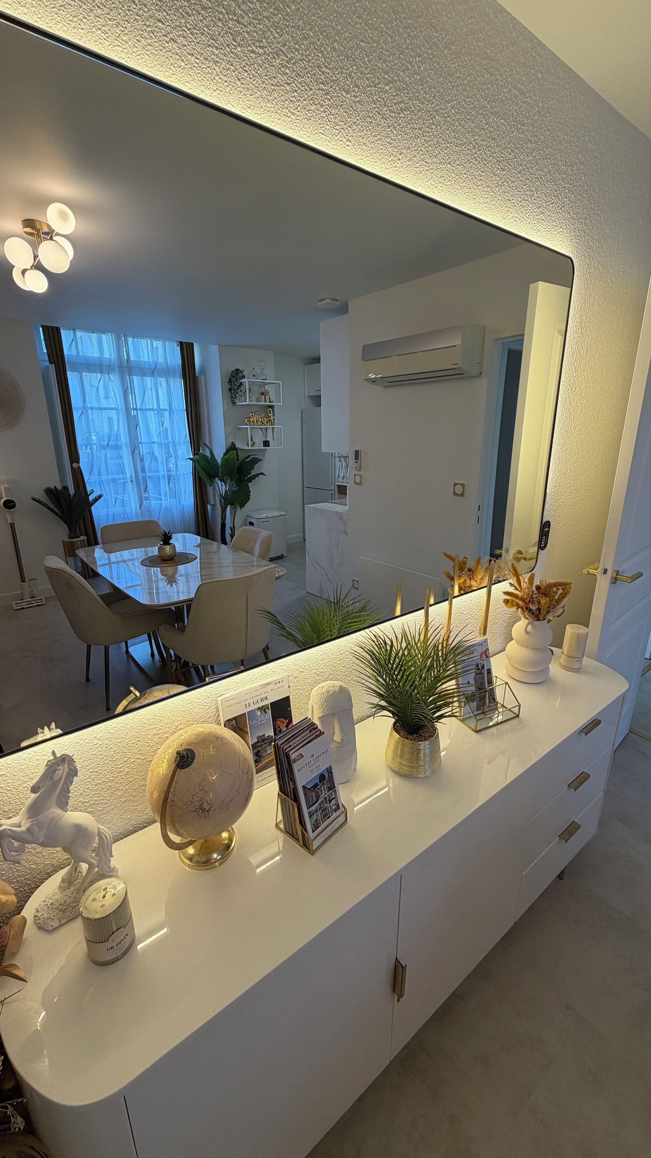 Un interno moderno con un tavolino consolle bianco, oggetti decorativi e un grande specchio che riflette una zona pranzo.