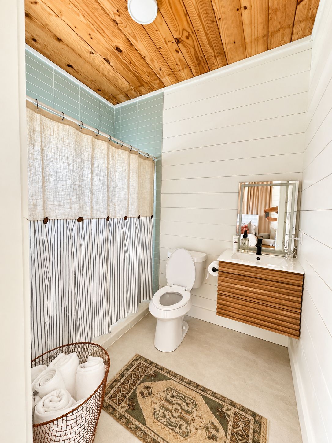 A bathroom with a wooden ceiling, a white toilet, a wooden vanity, a mirror, and a patterned rug.