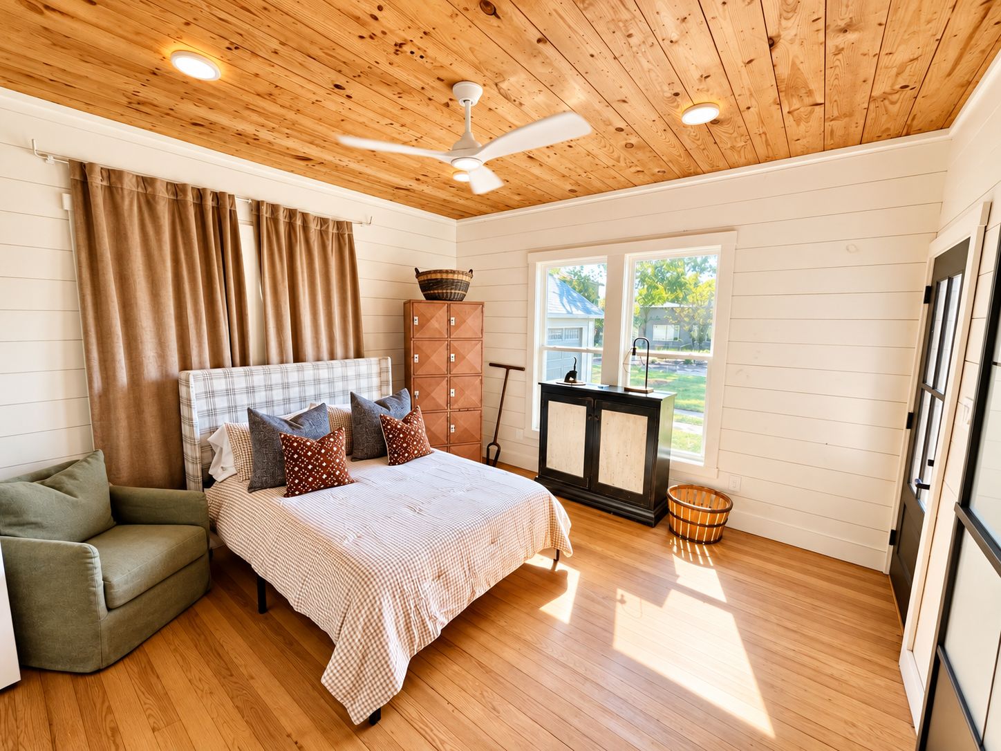 A bedroom with a bed, a green armchair, a wooden ceiling, and a window with a view of the outside.