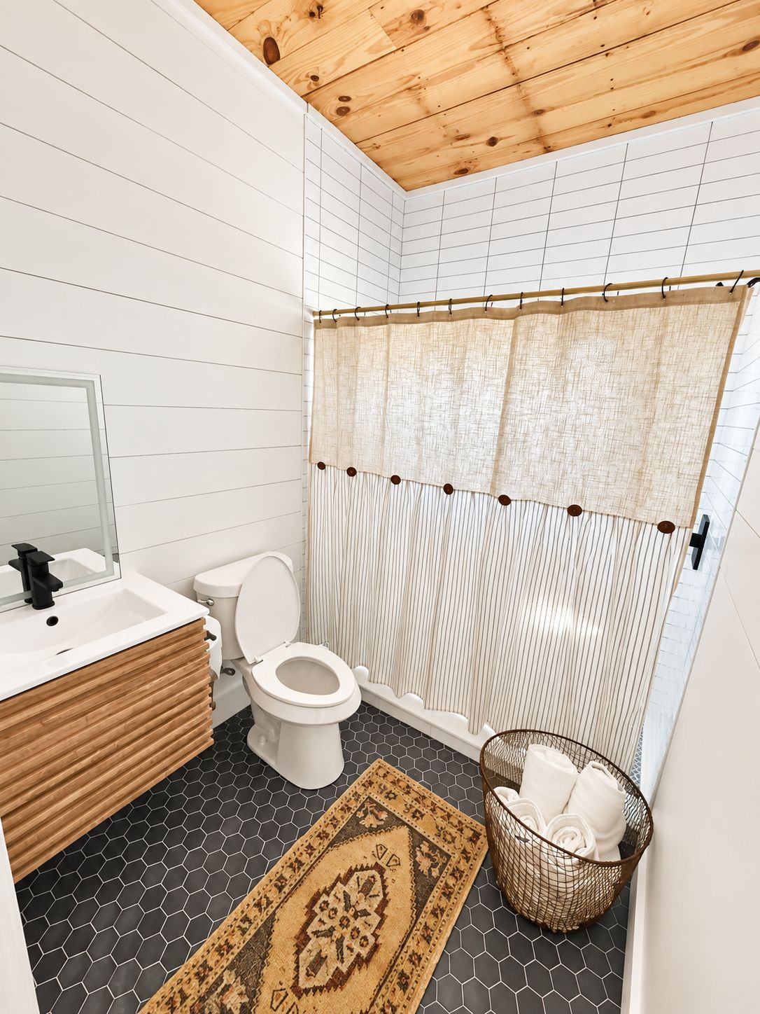 A bathroom with a wooden ceiling, white tiled walls, a white toilet, a sink with a mirror, and a shower curtain.
