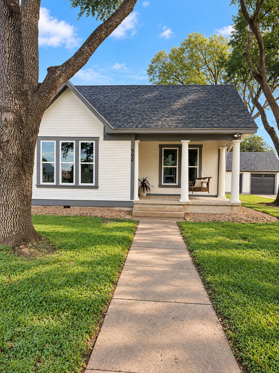 A single-story house with a front porch, a driveway, and a large tree to the left.