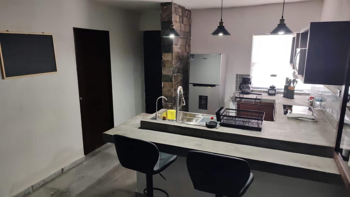 Una cocina moderna con una isla central, dos sillas negras, horno empotrado e iluminación colgante.