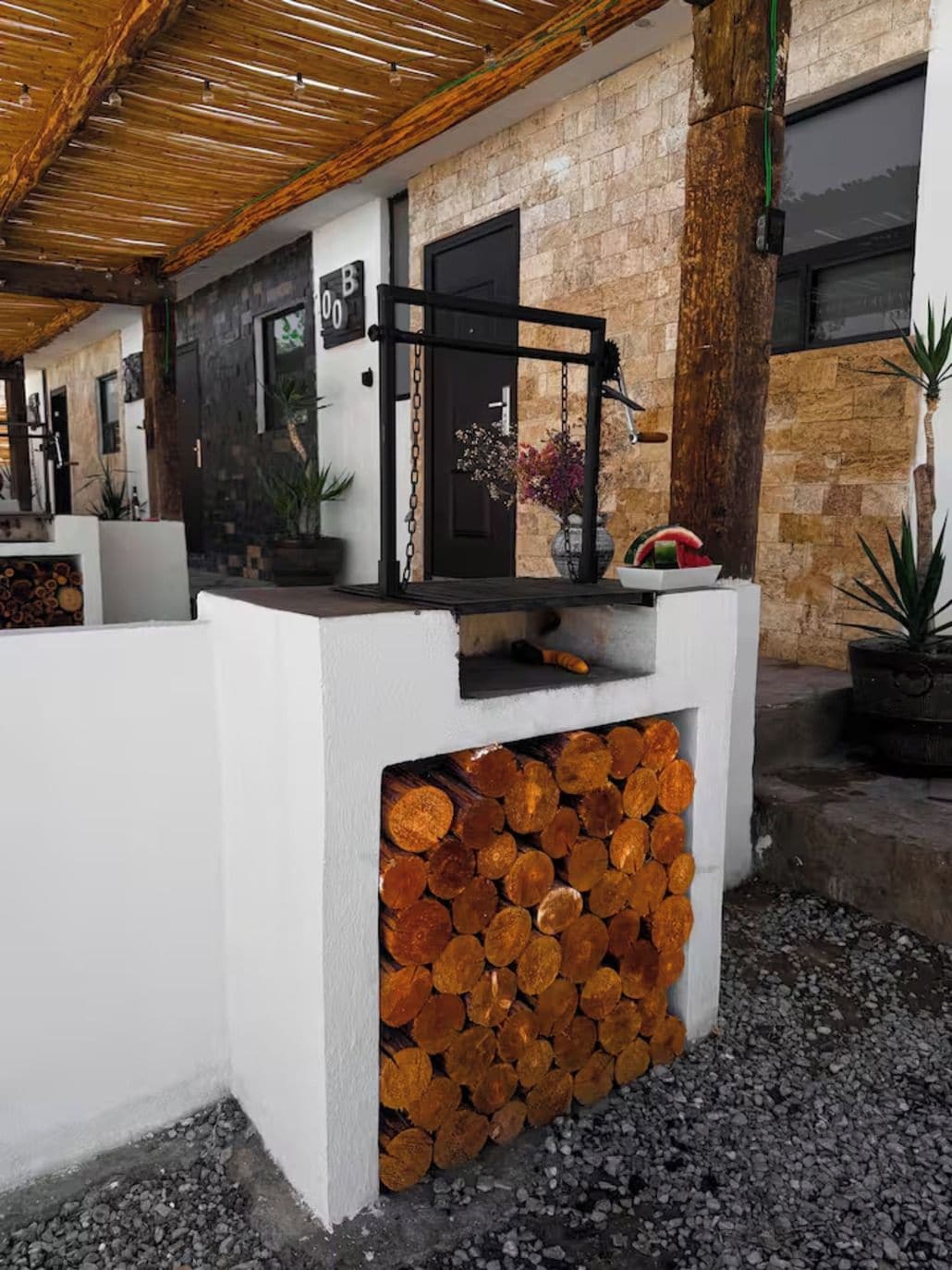Un área de estar moderna al aire libre, con una pila de leña, una puerta negra y un muro de piedra.