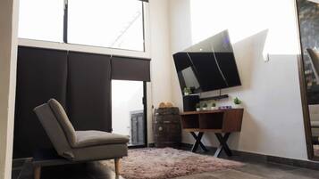A modern living room with a sofa, a flat-screen TV mounted on a wooden stand, and a large window providing natural light.