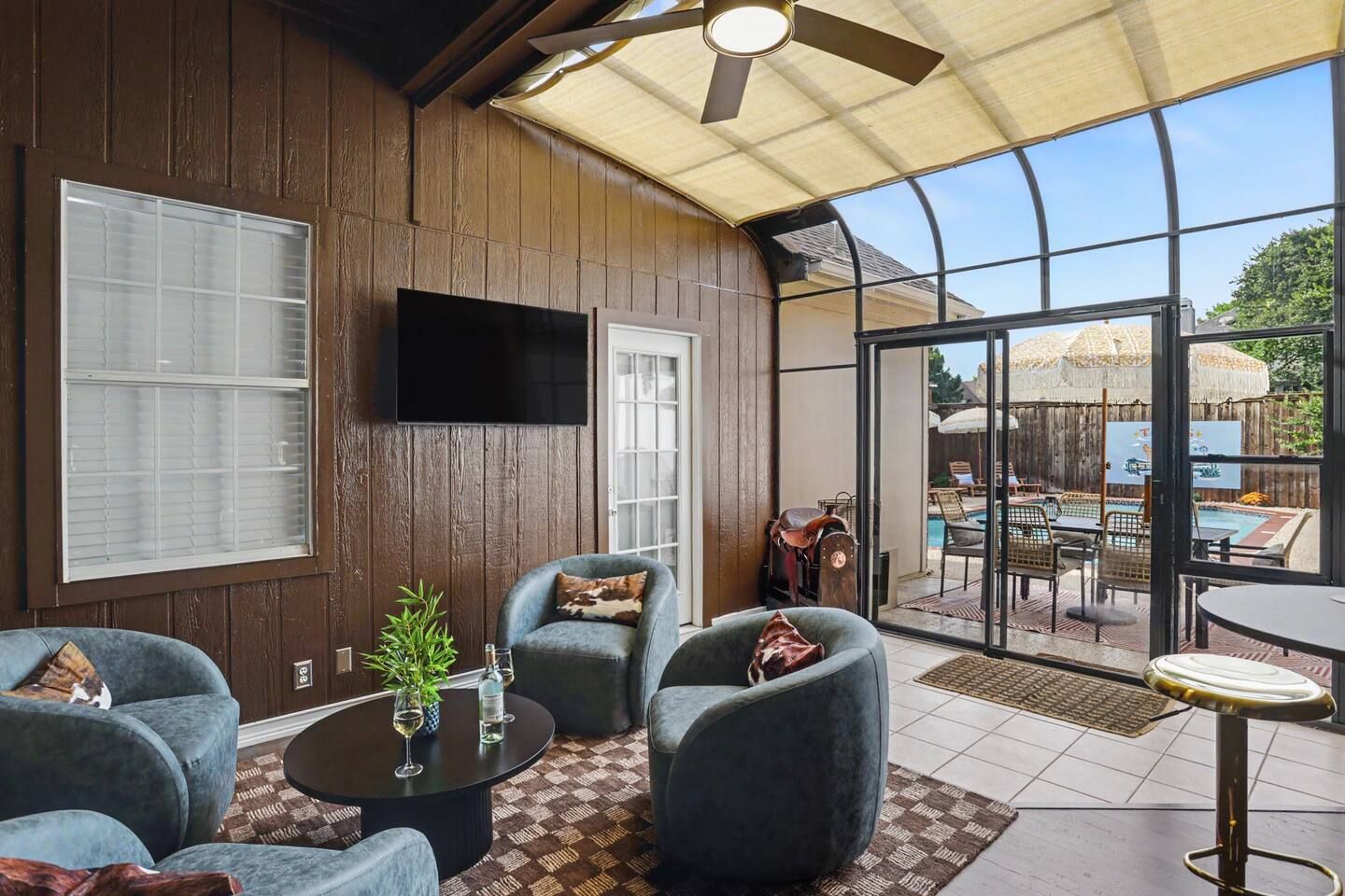 A sunroom with a ceiling fan, a flat-screen TV, and a view of an outdoor pool area.