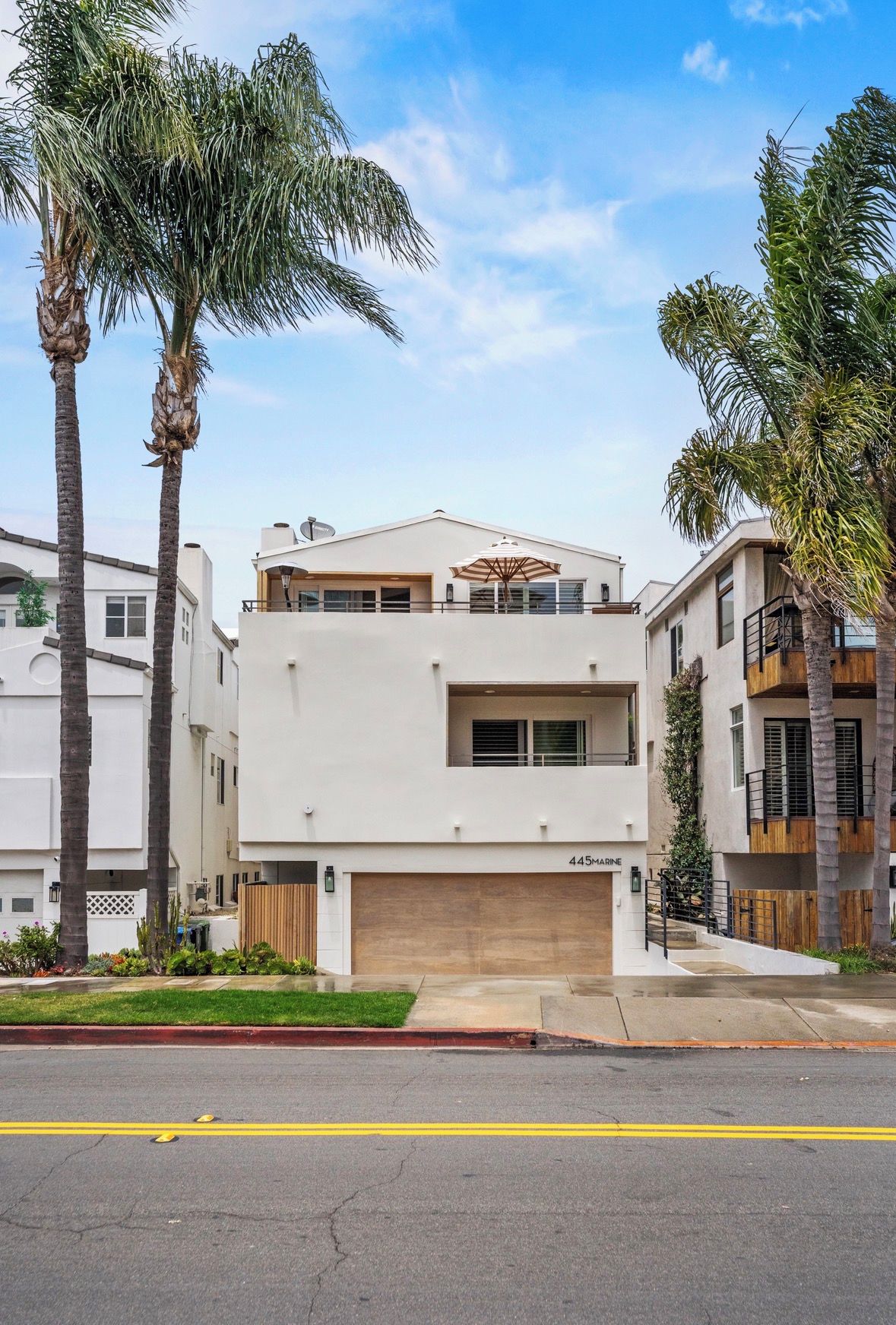 A modern two-story house with a garage, surrounded by palm trees and a clear sky.