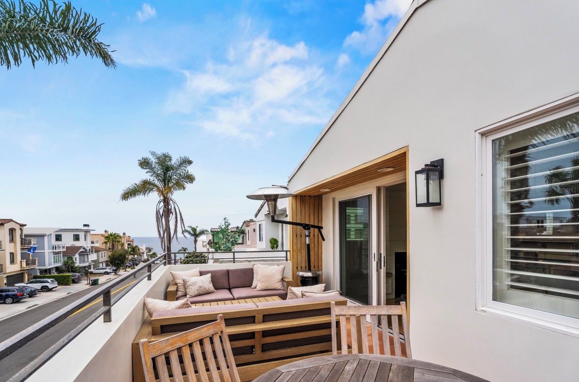 A balcony with a wooden bench, a sofa, and a table. There is a view of the street and palm trees in the background.