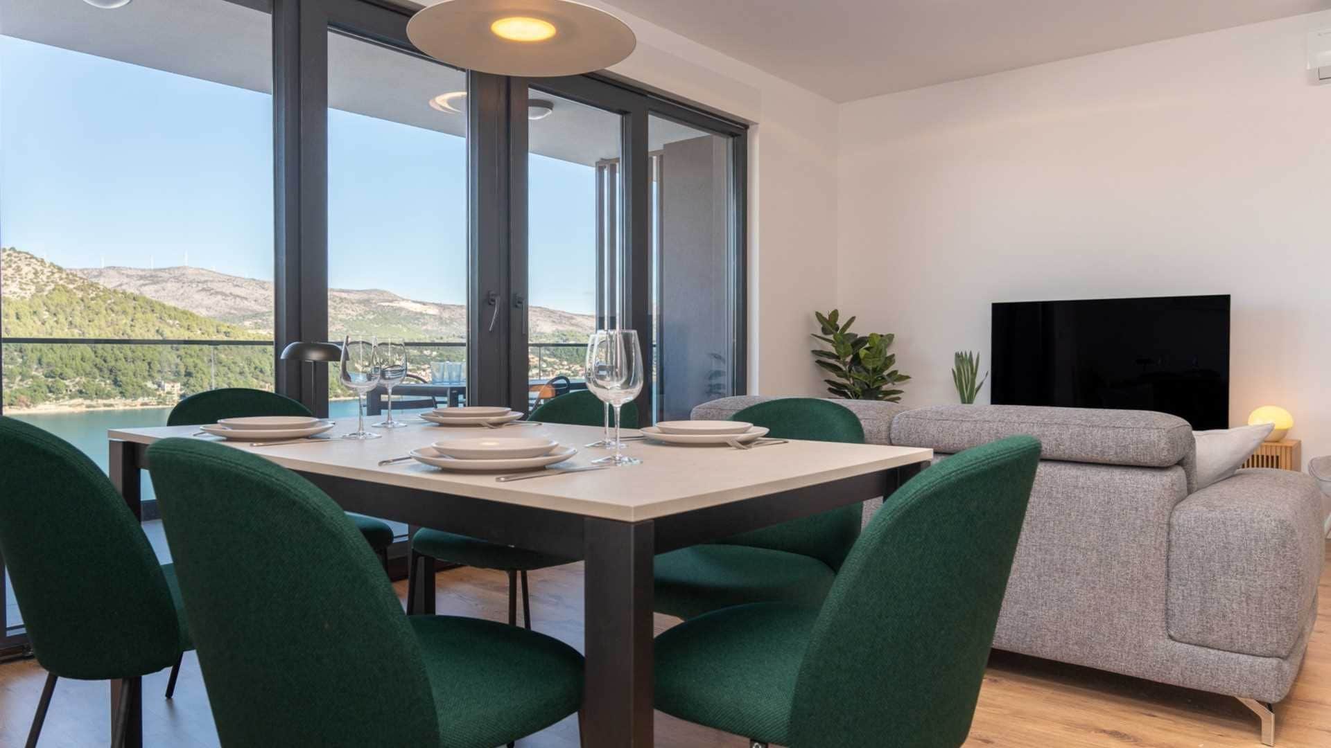 Comedor con mesa dispuesta para cuatro personas, ventanas amplias, sofá y televisor de pantalla plana.