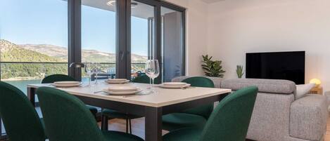 Comedor con mesa dispuesta para cuatro personas, ventanas amplias, sofá y televisor de pantalla plana.