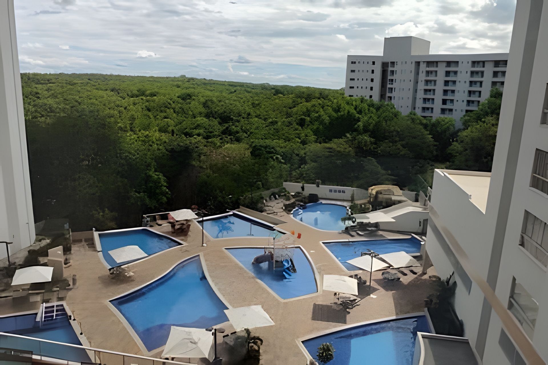Área de piscina no terraço com várias piscinas, cercada por vegetação e adjacente a um edifício de vários andares.