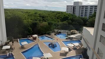 Área de piscina no terraço com várias piscinas, cercada por vegetação e adjacente a um edifício de vários andares.