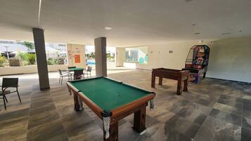 Espaço de lazer coberto com mesa de bilhar, mesa de futebol de mesa e área de estar com cadeiras e mesas.