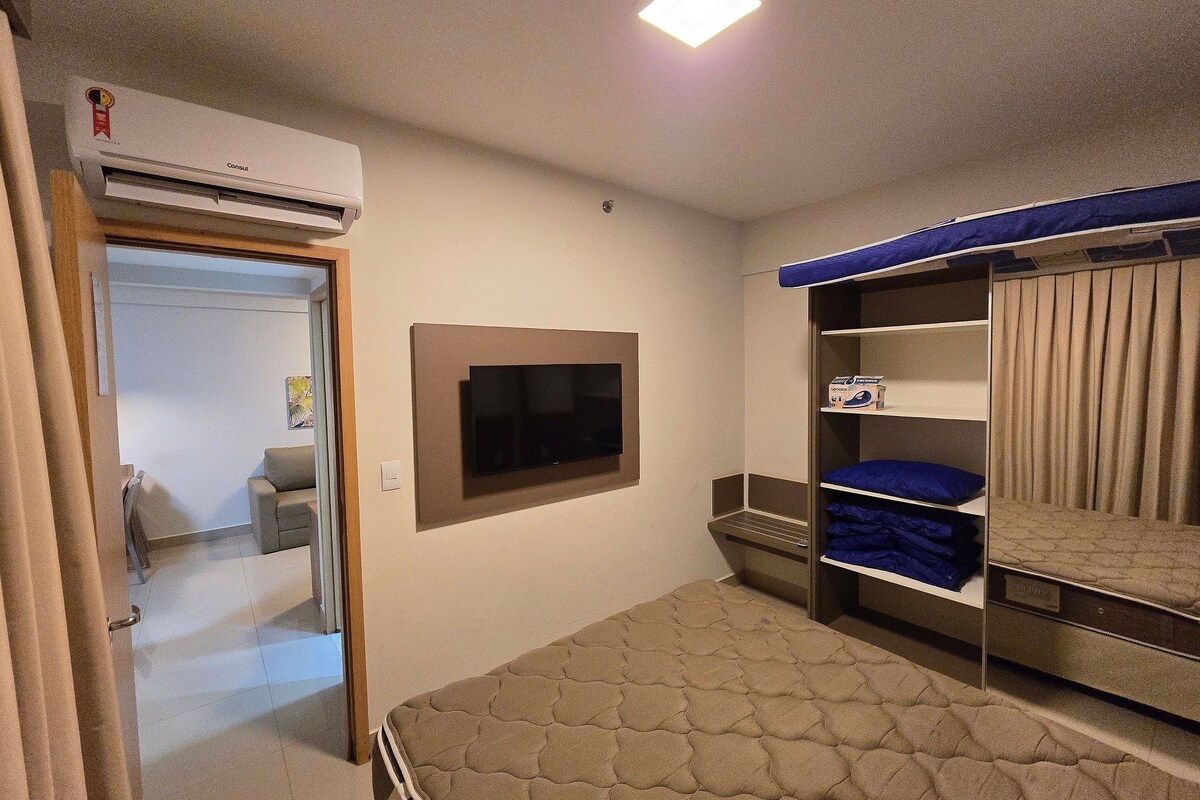 Una habitación con una cama, un televisor montado en la pared, un armario con repisas y un aire acondicionado.