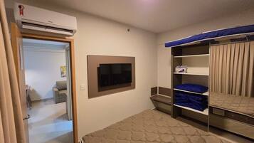 Una habitación con una cama, un televisor montado en la pared, un armario con repisas y un aire acondicionado.