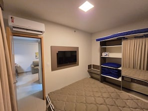 Una habitación con una cama, un televisor montado en la pared, un armario con repisas y un aire acondicionado.
