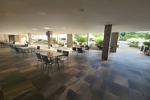 Un amplio espacio de comedor al aire libre con mesas y sillas, pilares grandes y vista a zonas verdes.