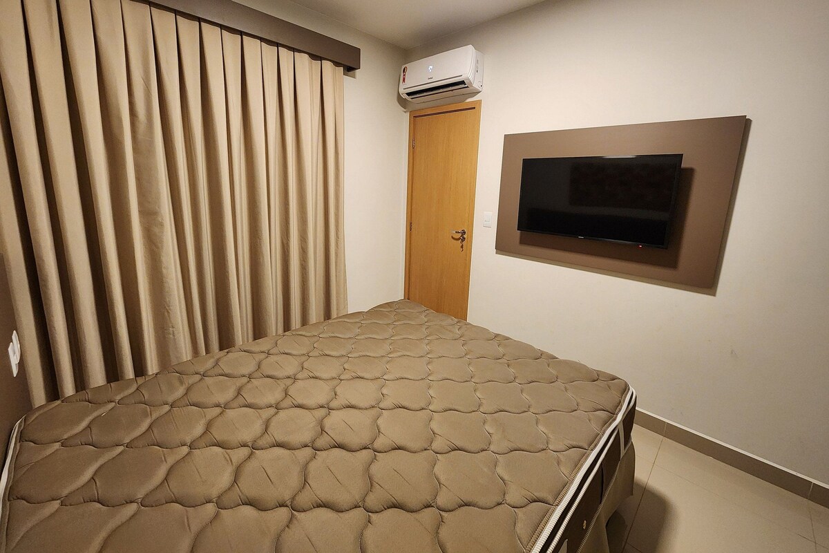 Una habitación con cama, unidad de aire acondicionado, puerta de madera y televisor montado.