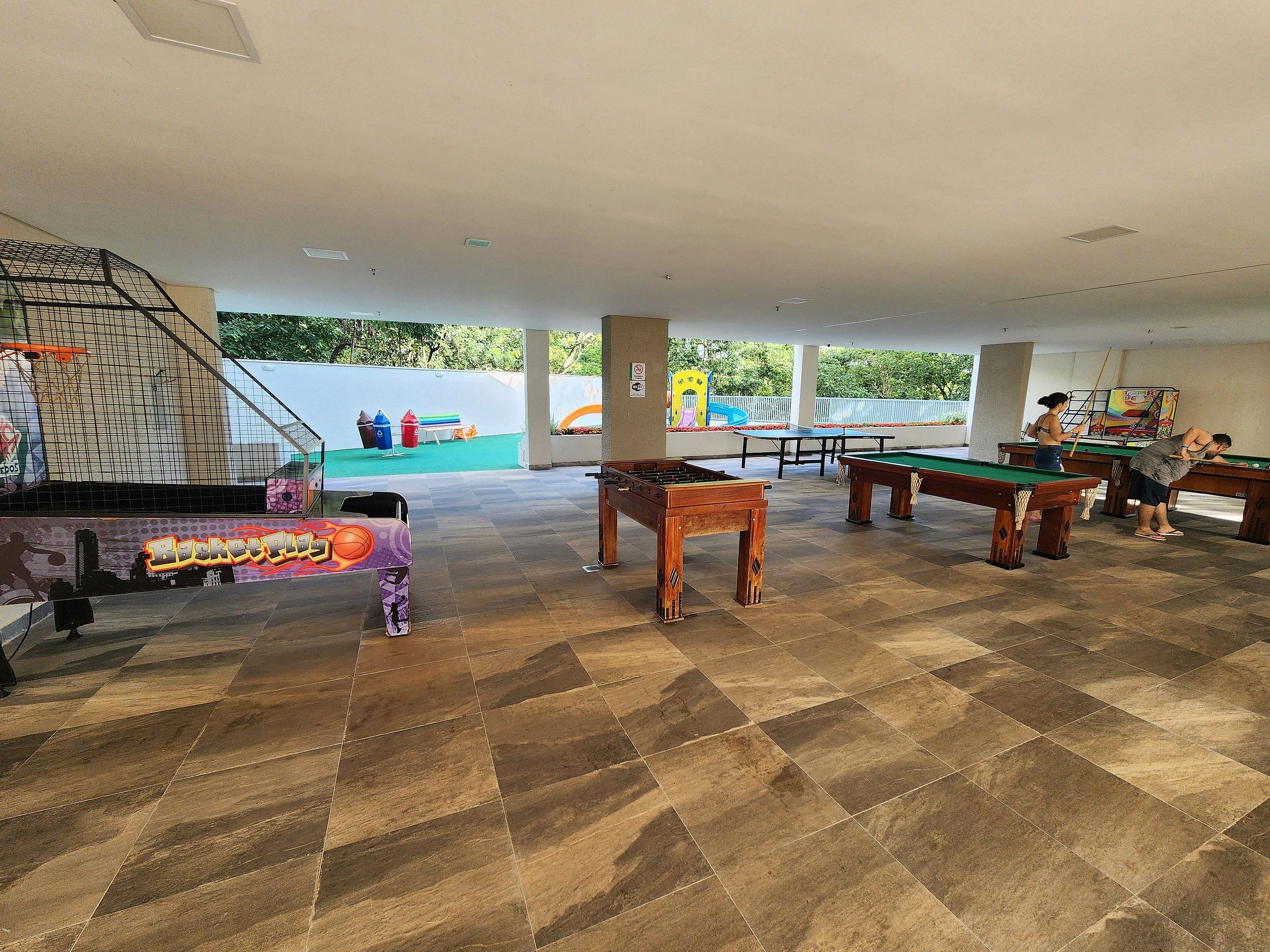 Un espacio recreativo interior con mesas de billar, una mesa de futbolín y una cancha de baloncesto.