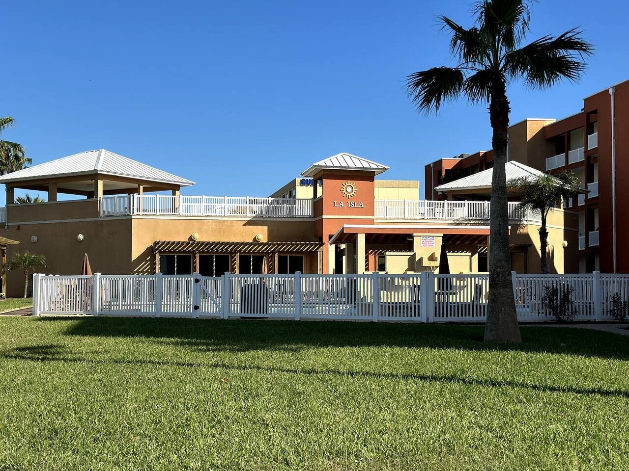 A hotel with a white fence, a palm tree, and a clear blue sky.