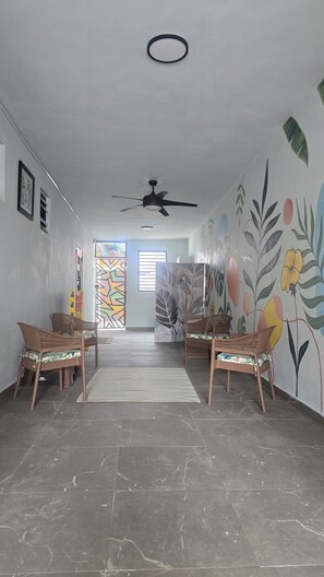 Amplia habitación con techo alto, ventilador de techo y ventana grande con vistas. Hay dos sillas de mimbre y una alfombra estampada en el suelo.