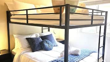 Ein Etagenbett mit Matratze und Kissen, ein Fenster mit Vorhängen sowie ein an der Wand befestigtes Regal mit zwei Kuscheltieren.
