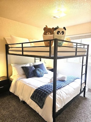 Ein Etagenbett mit Matratze und Kissen, ein Fenster mit Vorhängen sowie ein an der Wand befestigtes Regal mit zwei Kuscheltieren.
