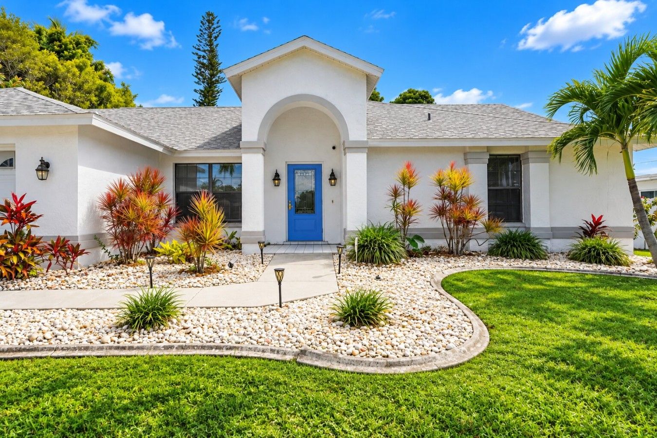 A two-story house with a blue front door, white exterior walls, and a landscaped front yard with various plants and a gravel pathway.