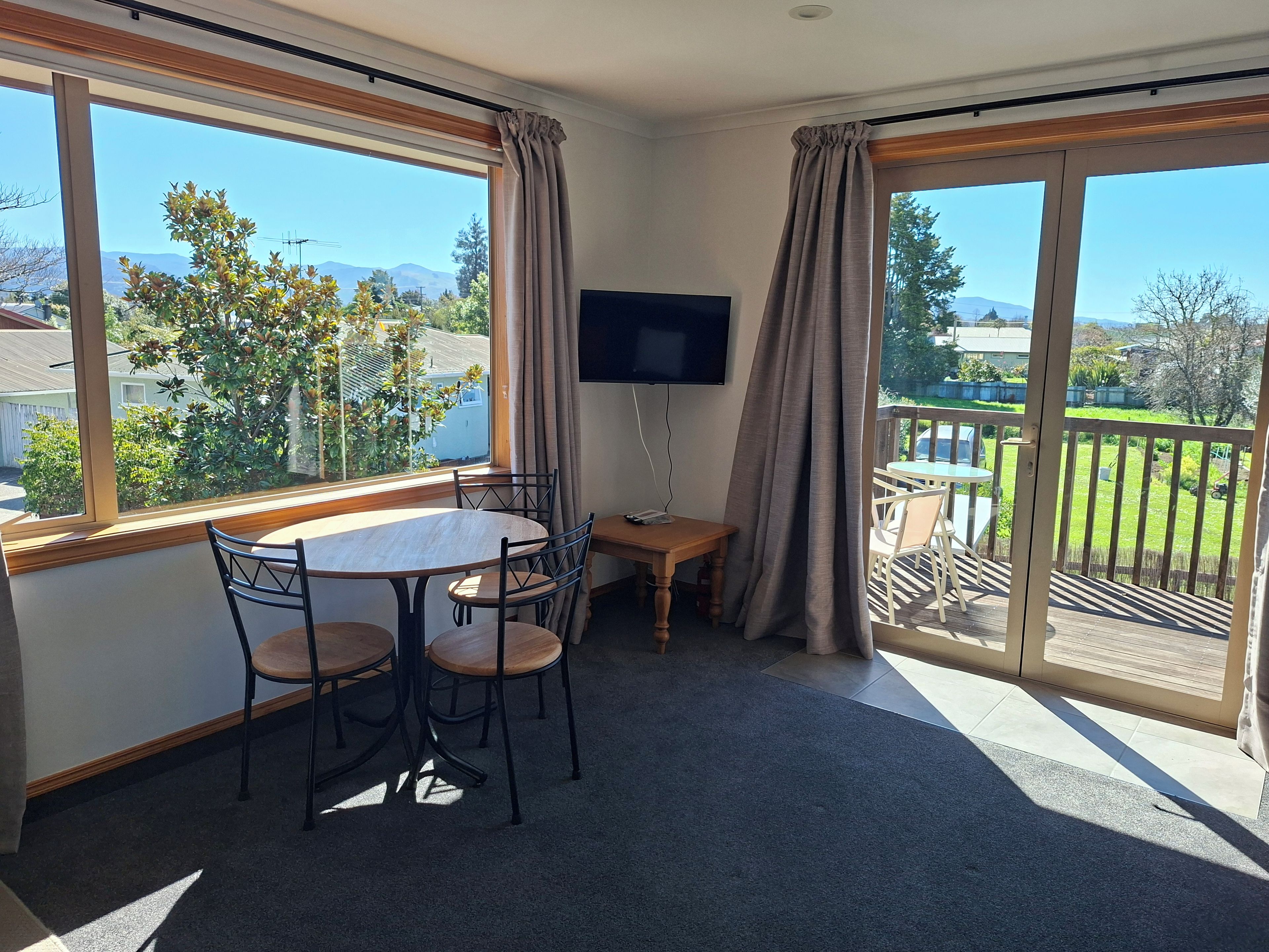 A room with a round table and chairs, a flat-screen TV mounted on the wall, and large windows with curtains.