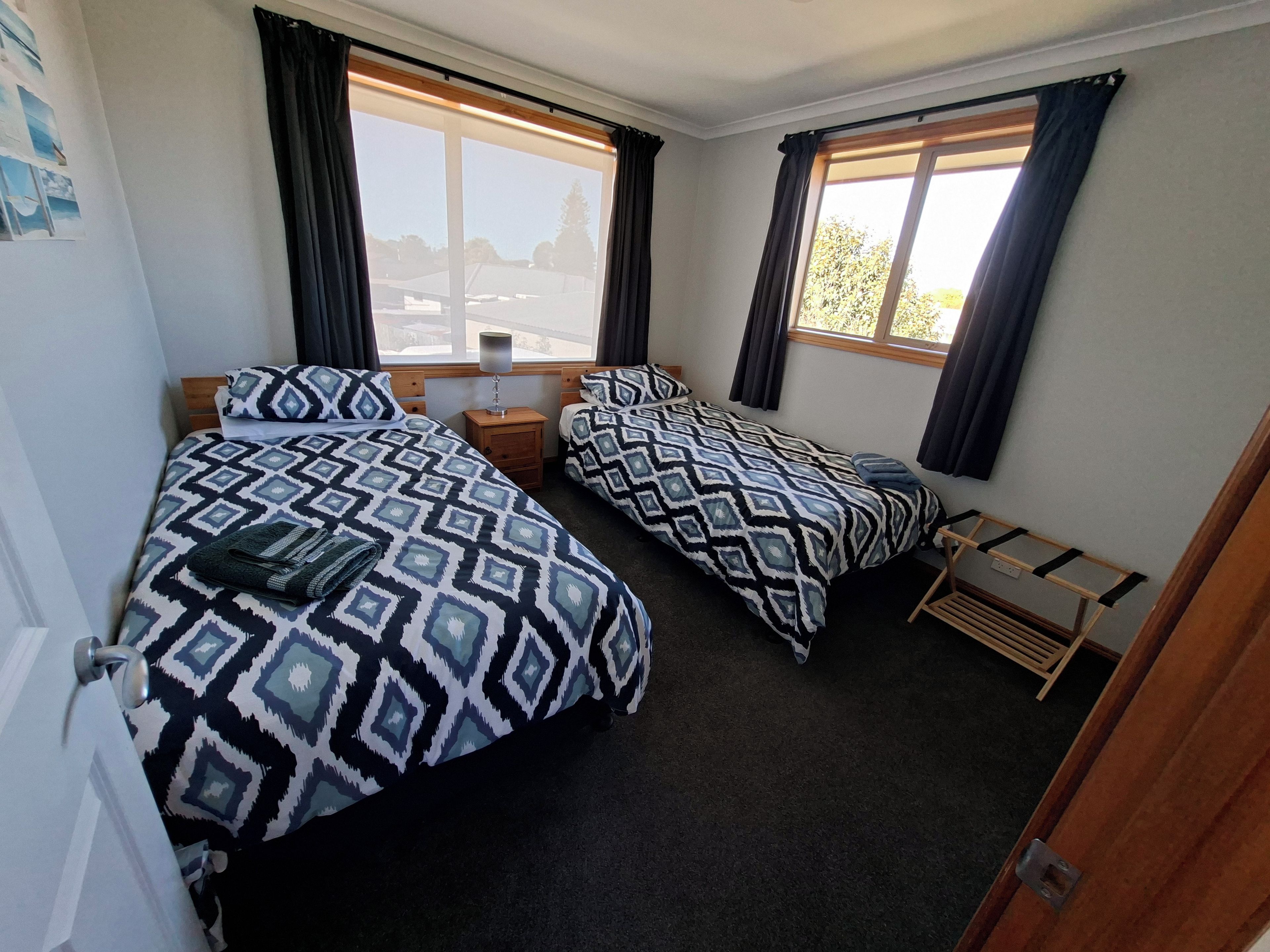 Quarto com duas camas, mesa de cabeceira, janela com cortinas e prateleira de madeira.