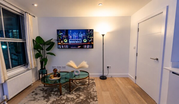 En moderne stue med sofabord i glas, et fladskærms-tv ophængt på væggen, en gulvlampe og en dør.