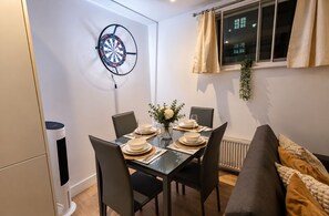 Et spiseområde med et bord dækket til fire personer, en dartskive, en radiator og et vindue med gardiner.