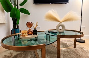 A modern living room with a glass coffee table, a round side table, a potted plant, decorative items, and a television mounted on the wall. - Stylish 2-Bed Apartment -Canary Wharf - Sleeps 5- Near City Airport & Westfields (London)