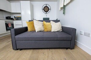 En grå sofa med fire puder i en moderne stue, hvor et køkken ses i baggrunden.